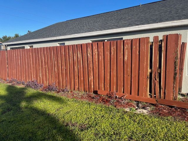 Red wooden fence in disrepair, with some panels missing, against a house and sunny sky.