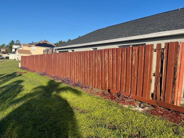 Brown wooden fence alongside a green lawn, next to a house with a black roof under a blue sky.