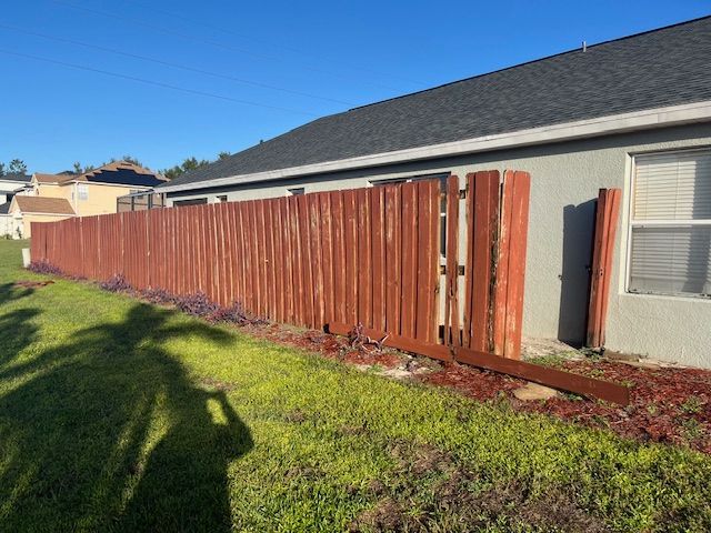 Red wooden fence along a gray house with a grassy yard and blue sky.