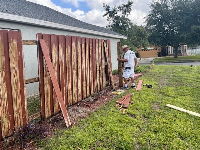 A person repairs a red wooden fence on a sunny day, with spare boards nearby.
