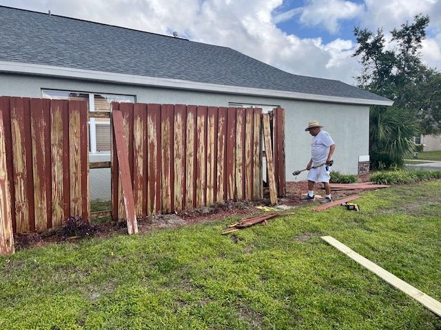 Man repairing weathered red wooden fence next to a house on a cloudy day.