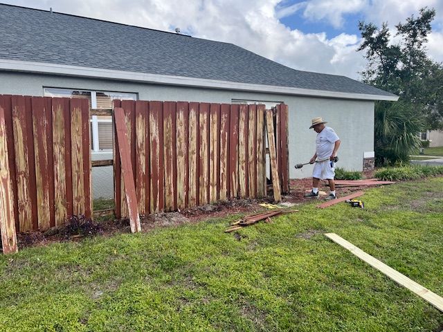 Man paints a weathered red wooden fence next to a pale house with a gray roof; grass and blue sky.