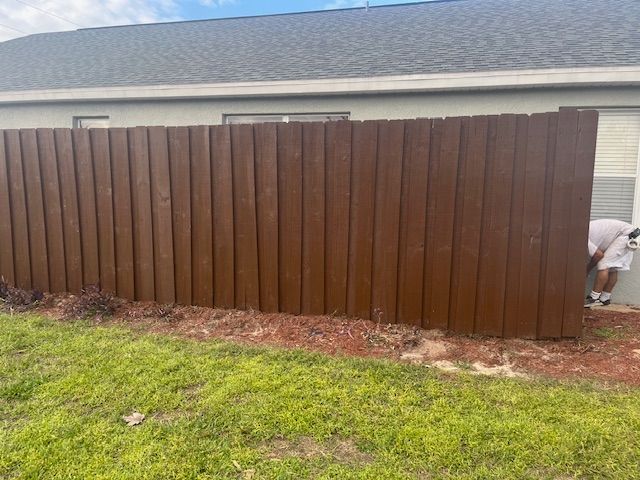 Brown wooden fence against a building; person in white working on right side; green grass and mulch below.