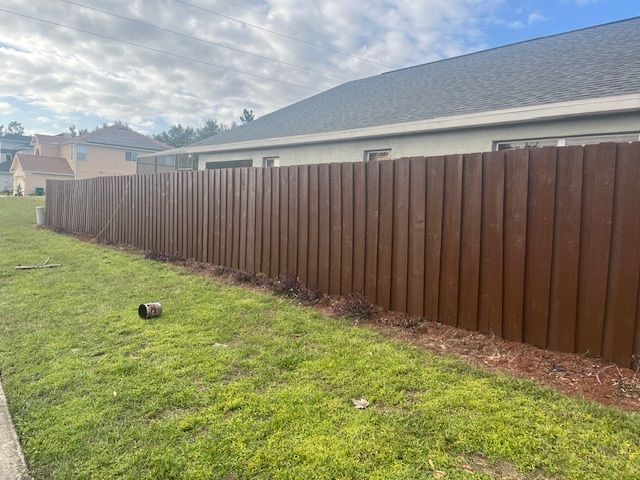 Brown wooden fence along a grassy yard, with a house in the background under a cloudy sky.
