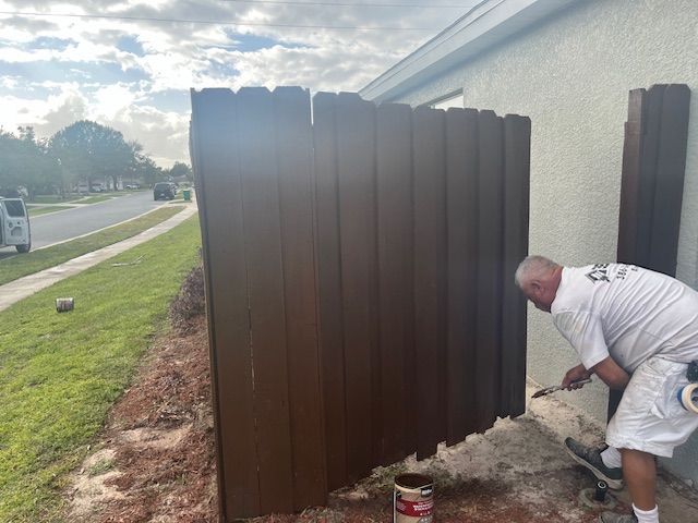 Man paints a brown wooden fence next to a house, outdoors on a sunny day.