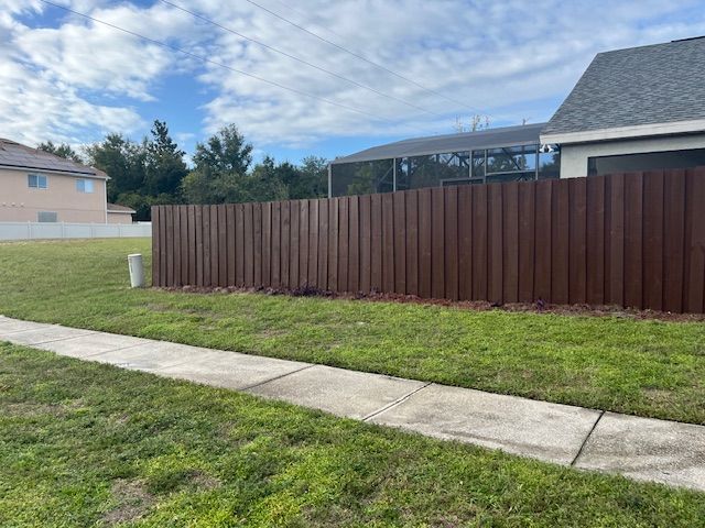 Brown wooden fence borders a green lawn and a concrete sidewalk next to a house.