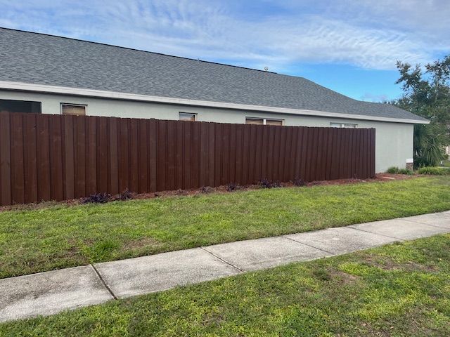 Brown wooden fence along a house with green grass and a sidewalk.