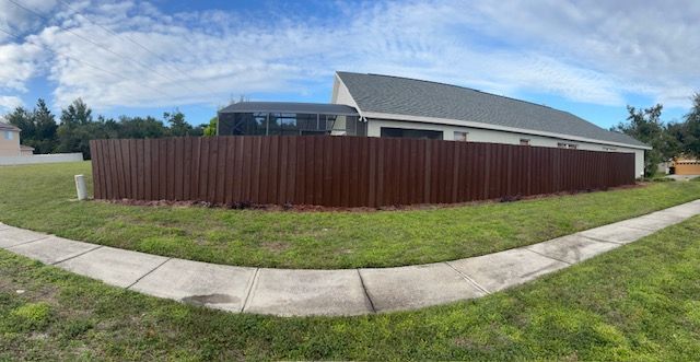 Brown wooden fence surrounds a house with a gray roof and lawn on a sunny day.