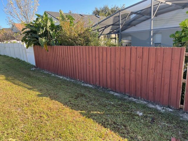 Red wooden fence in a yard with patches of grass, with a white fence section to the left.