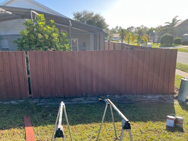 A red-brown stained wooden fence in a backyard with a sunny sky. Sawhorses hold paint cans.