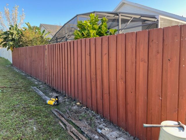 Red wooden fence in a backyard, a lawnmower and gardening tools on the ground, sunny day.