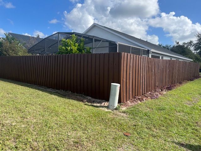 Brown wooden fence surrounding a house with a screened enclosure, set in a grassy yard under a partly cloudy sky.