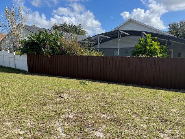 Brown and white fences in a grassy yard, with a house and a screened pool in the background under a partly cloudy sky.