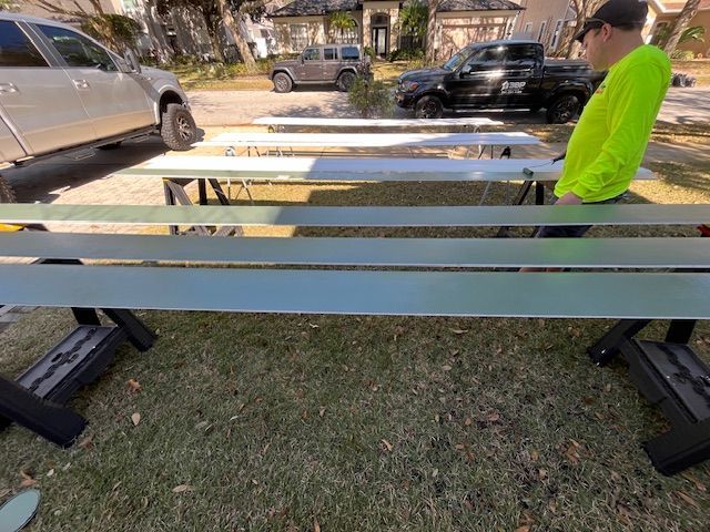Man in neon shirt inspects metal beams on a grass lawn, with trucks and a house visible.