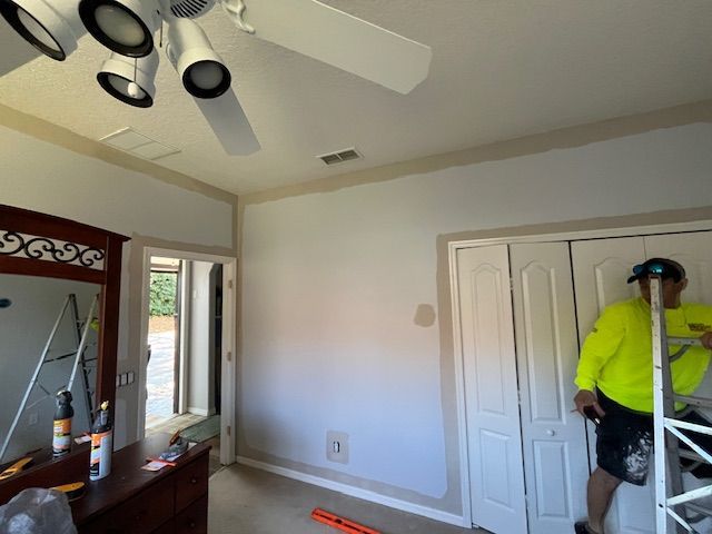 Man painting trim in bedroom with ceiling fan, closet and dresser.