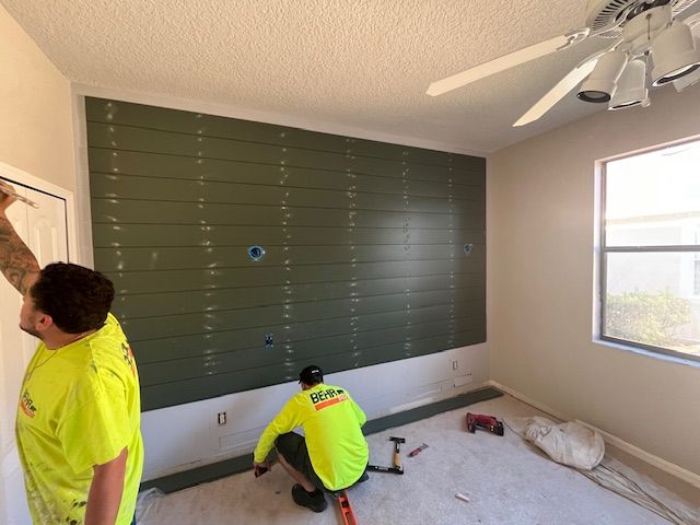 Two workers install a green shiplap accent wall in a room. One measures, the other kneels to work.