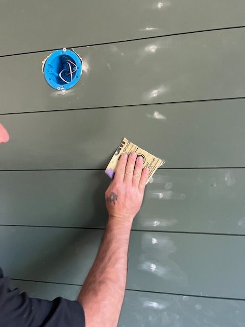 A person sands a dark green, horizontal wall with white patching in the sunlight.