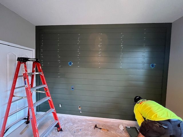 A person installs dark green shiplap wall, a red ladder stands nearby.