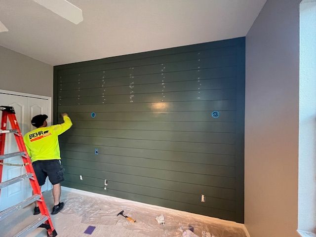 Person painting a dark green shiplap accent wall in a room; red ladder, white door, beige walls.