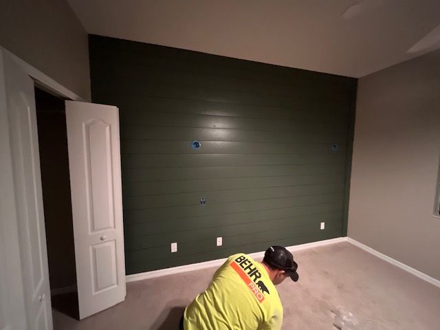 Man painting an accent wall dark green. A white door and beige carpet are visible.