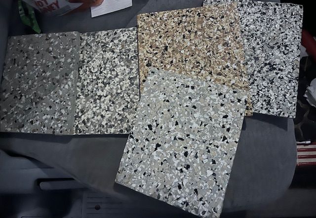 Five speckled floor tile samples in varying shades of gray, beige, and black, laid on a gray surface.