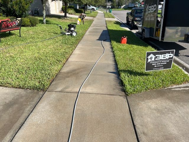 A worker pressure washes a concrete sidewalk, with a lawnmower and trailer visible.