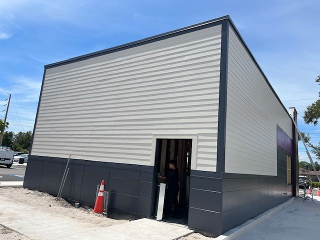 Exterior of a building under construction with gray and white siding, a partially open doorway, and concrete pathway.
