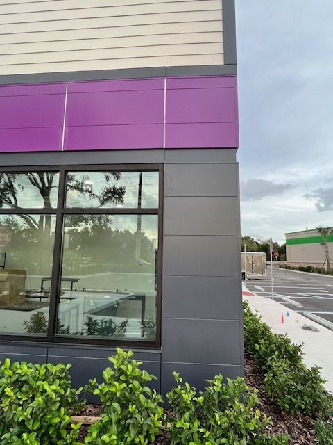 Exterior corner of a building with purple and gray panels, large window, and landscaping.