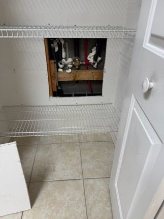 Inside a utility closet, a rectangular opening reveals plumbing and a wooden beam. White shelves and tile flooring present.