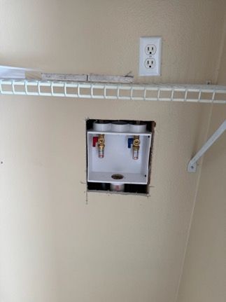 White washing machine water supply box with red and blue valves, installed in a wall. Electrical outlet and shelf visible.
