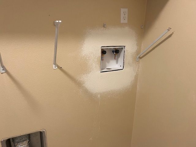 Laundry room wall with a water hookup box and shelf brackets; painted patch around the box.