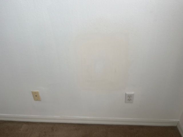 White wall with a discolored square, two electrical outlets, and a brown carpet.