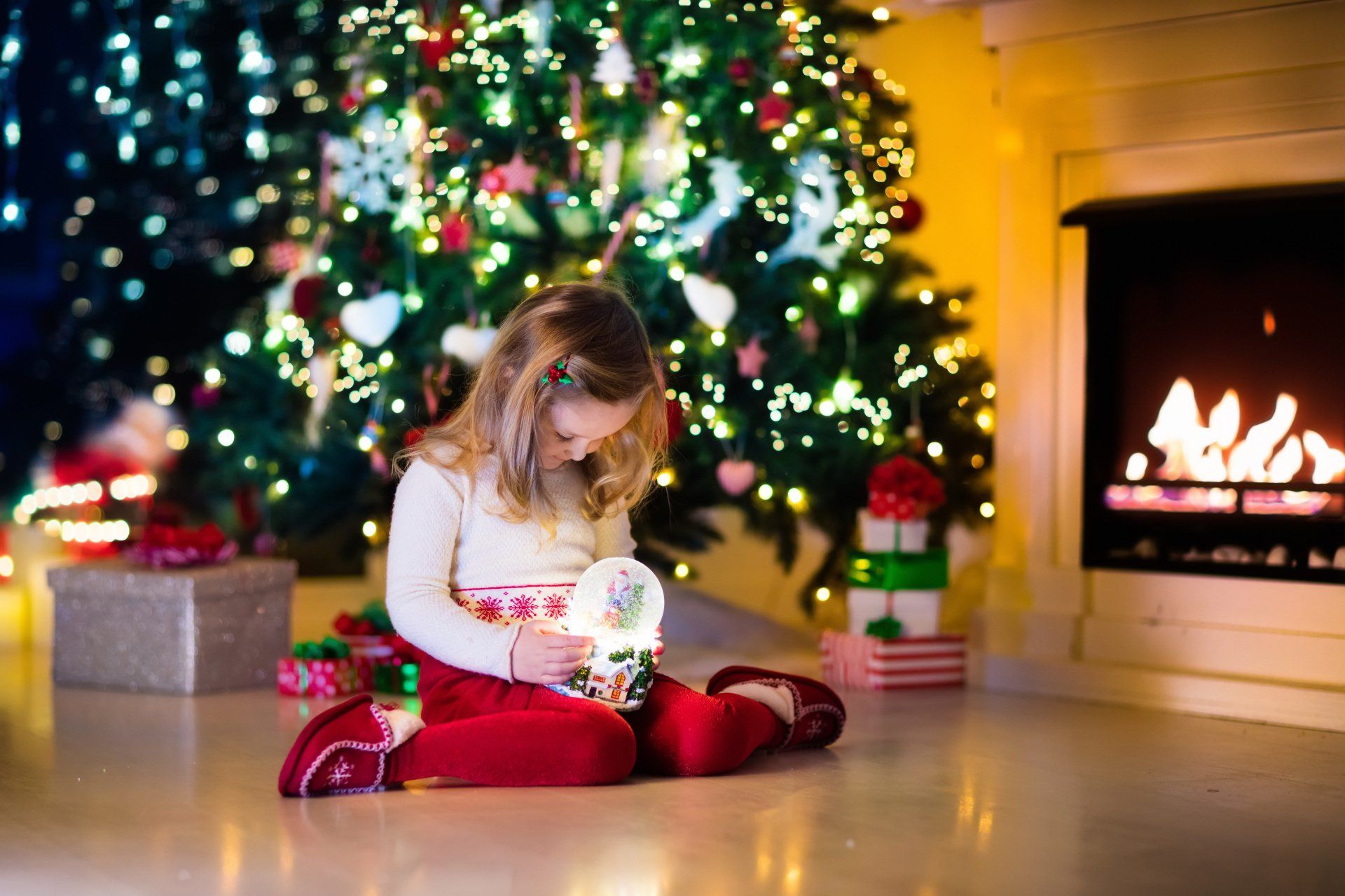 A little girl sits on the floor looking down at a snow globe with a Christmas tree in the background