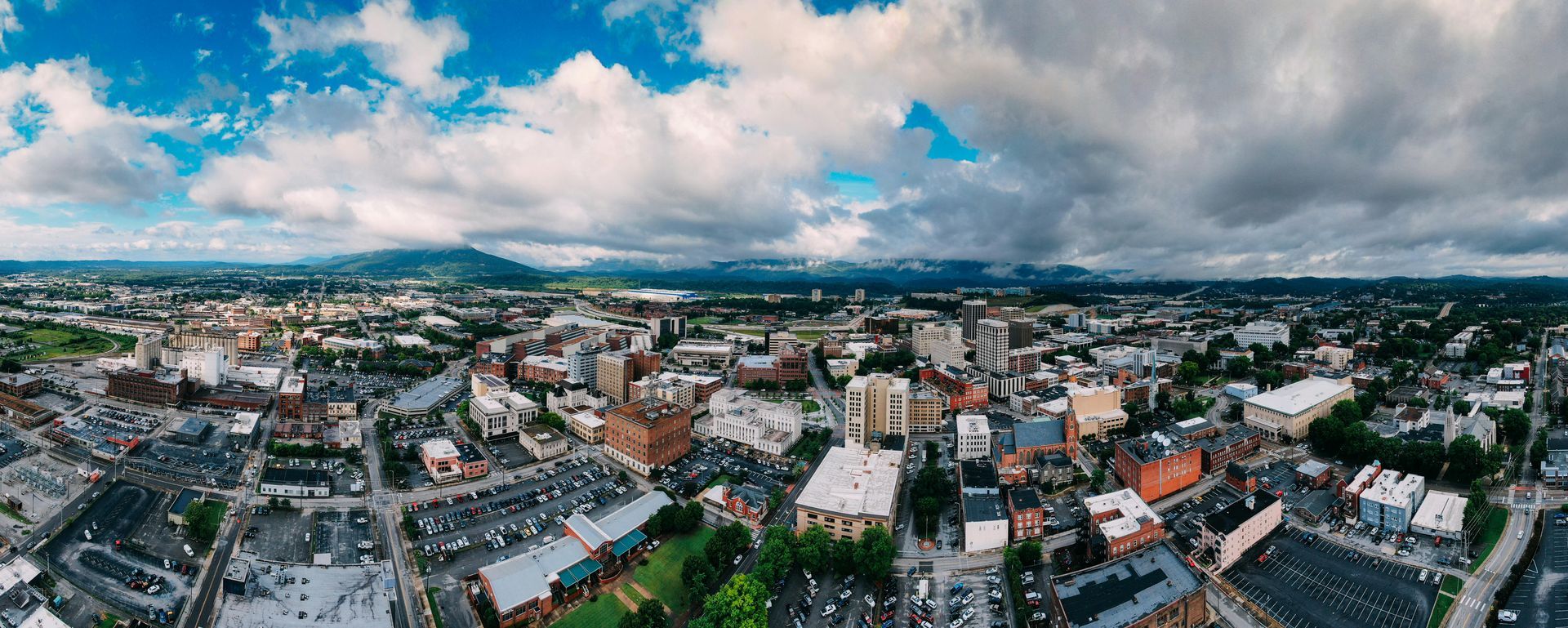 Panoramic aerial view of a city under a cloudy sky. Buildings, roads, and trees fill the urban landscape with a mountain range in the distance.