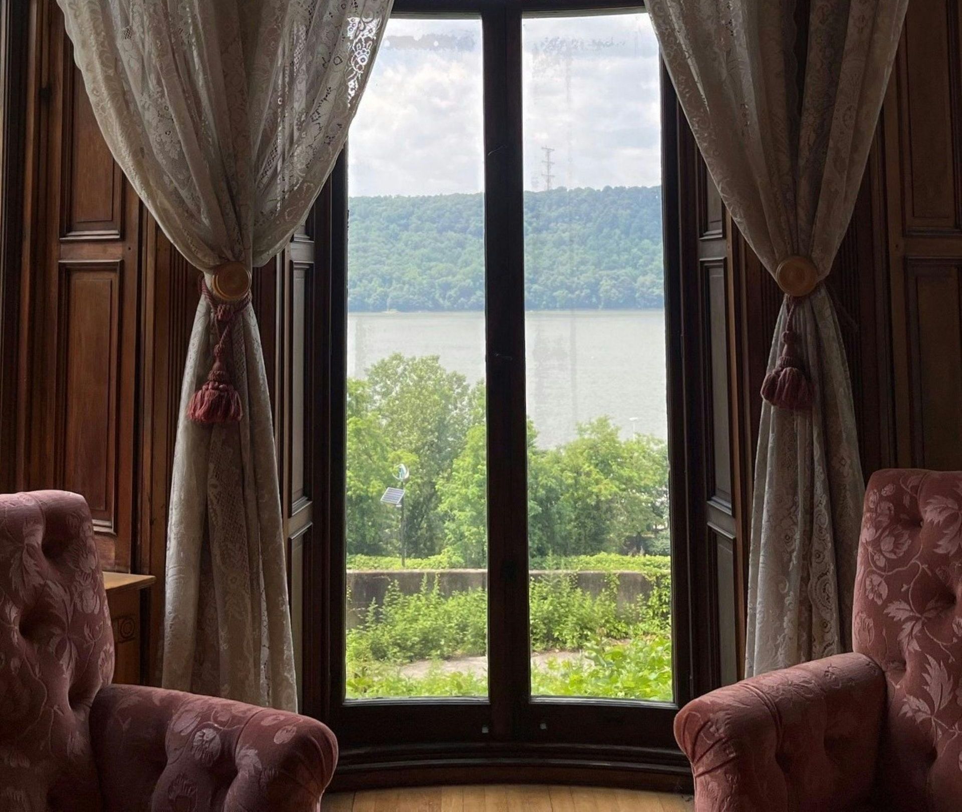 View through a window: river, trees, and sky seen from inside a room with wood paneling, lace curtains, and pink armchairs.