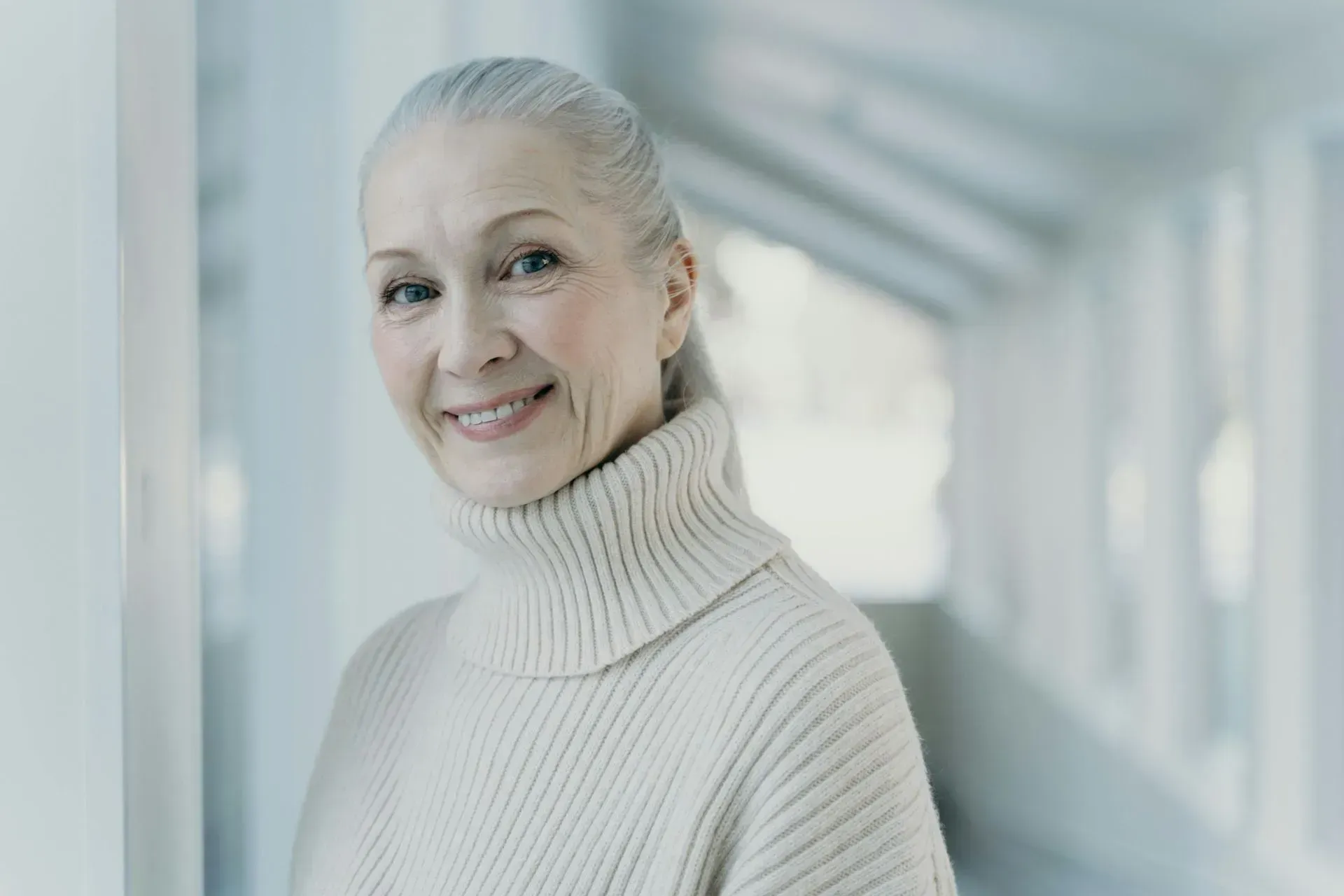 An elderly woman wearing a white sweater is leaning against a wall and smiling.