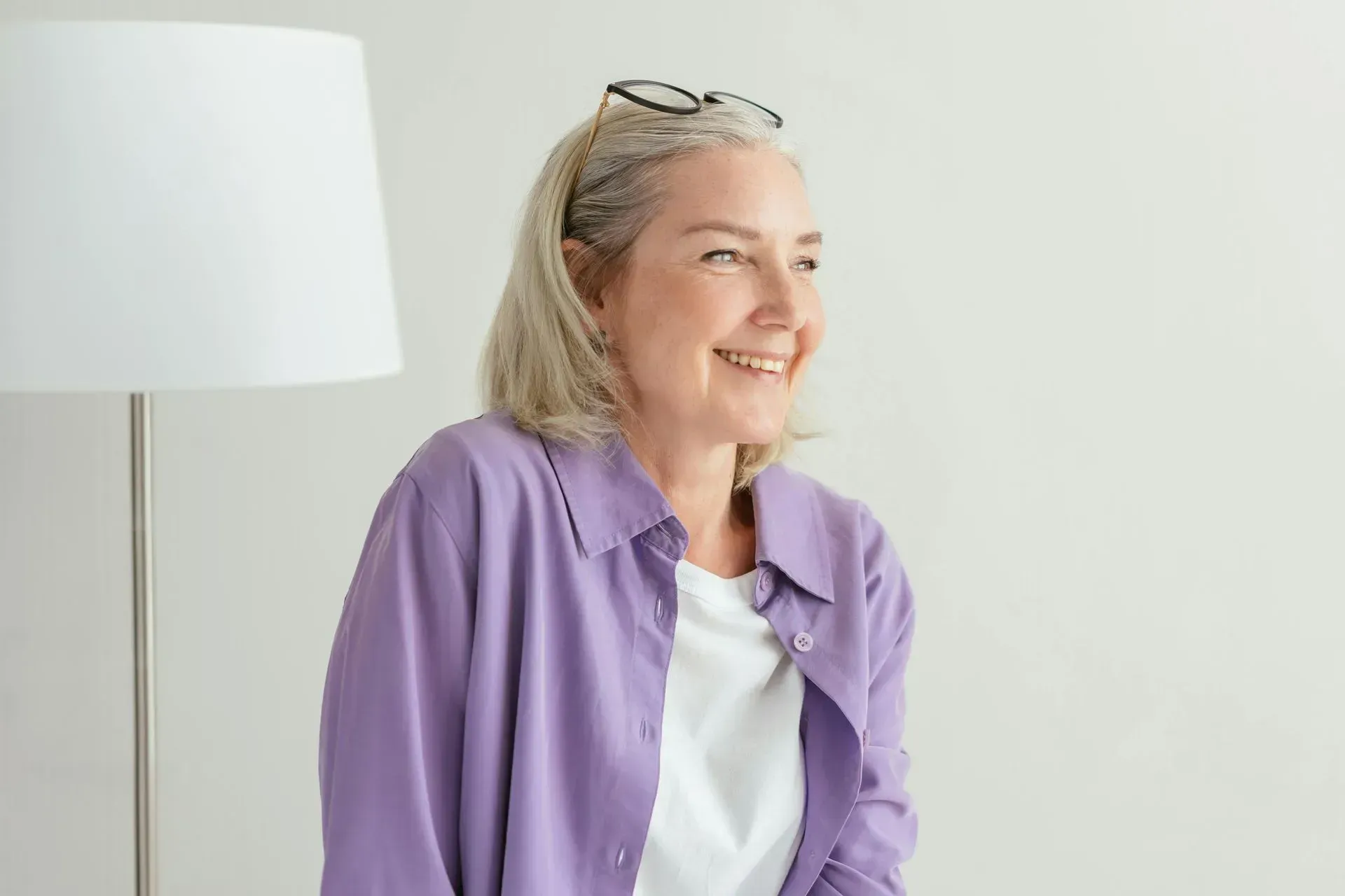 A woman in a purple shirt and glasses is smiling while sitting in front of a lamp.