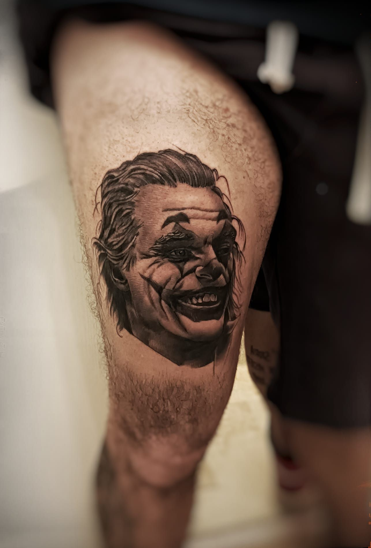 Tatuaggio del volto del Joker sulla coscia di una persona; ombreggiatura in bianco e nero, espressione sorridente.