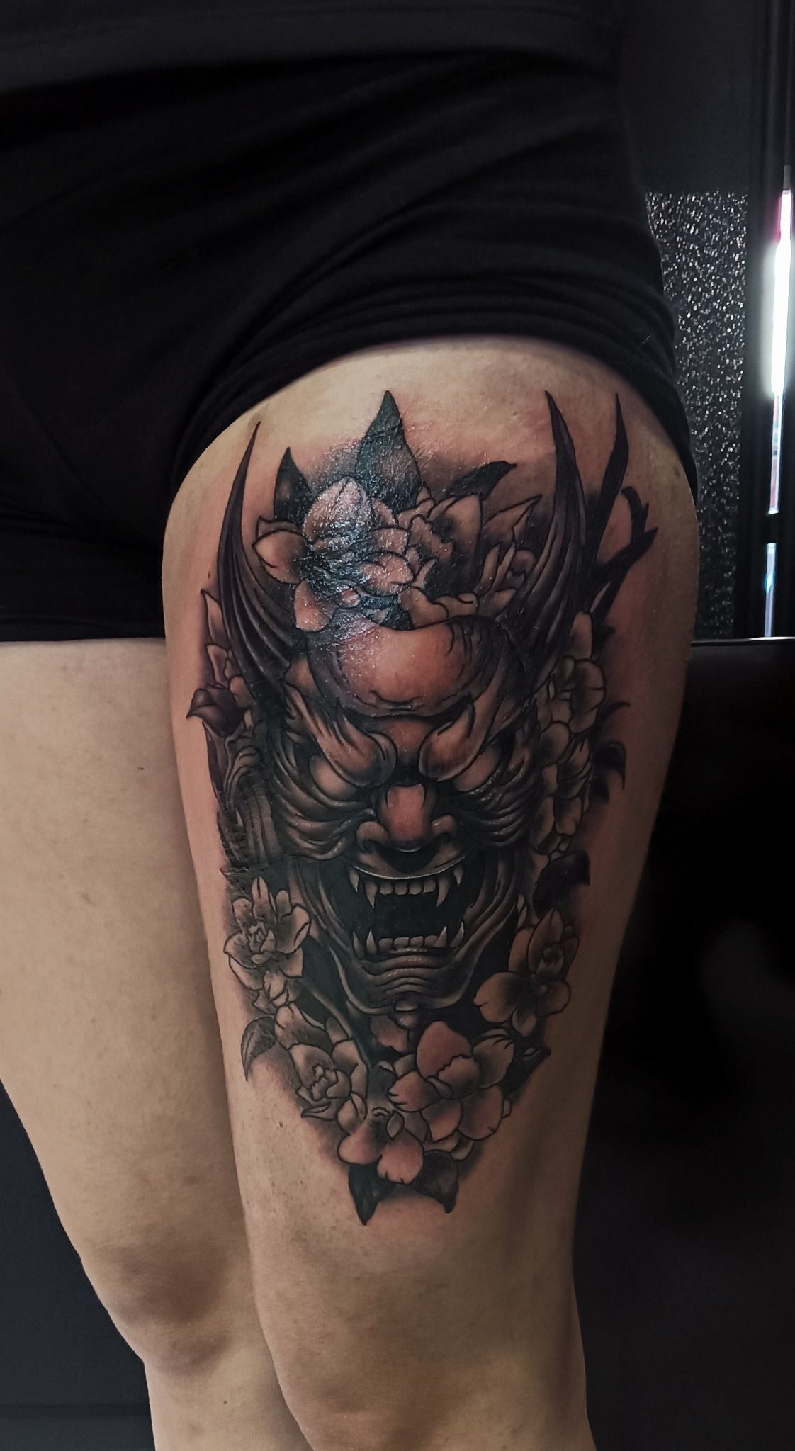 Tatuaggio di una maschera Oni nera e grigia con dettagli floreali sulla coscia di una persona.
