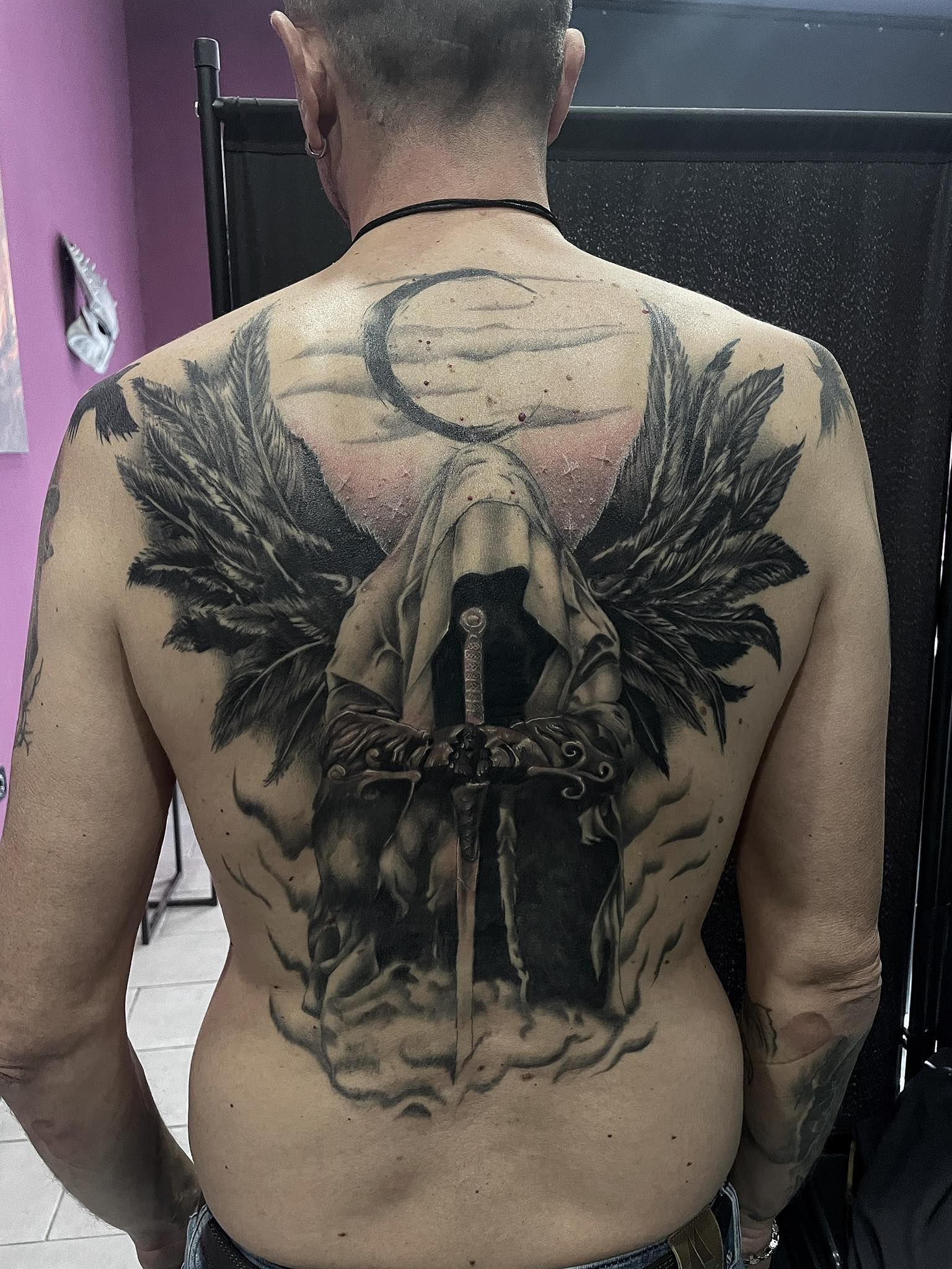 Schiena di un uomo con un grande tatuaggio: una figura ammantata, con ali e spada, sullo sfondo di un cielo illuminato dalla luna.