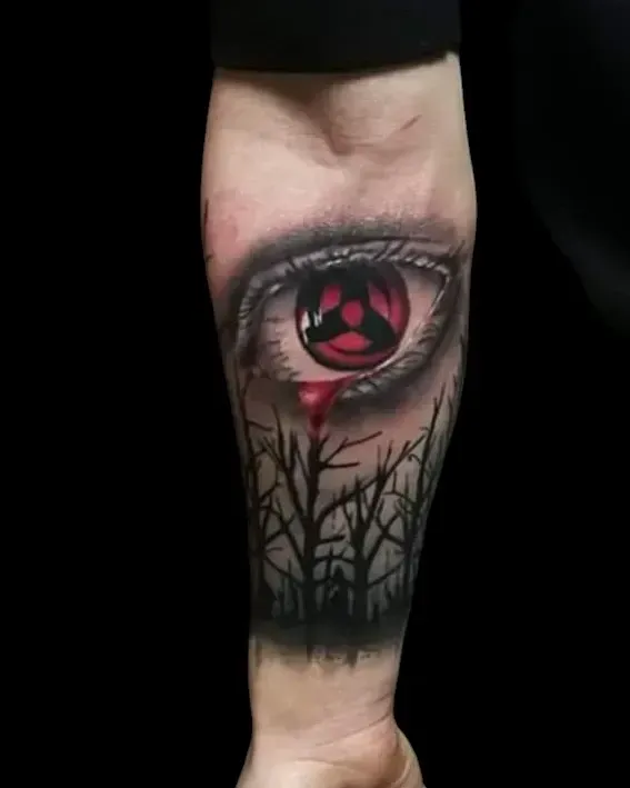 Tatuaggio di un occhio Sharingan con lacrima di sangue e alberi in silhouette sull'avambraccio. Rosso, nero e grigio.