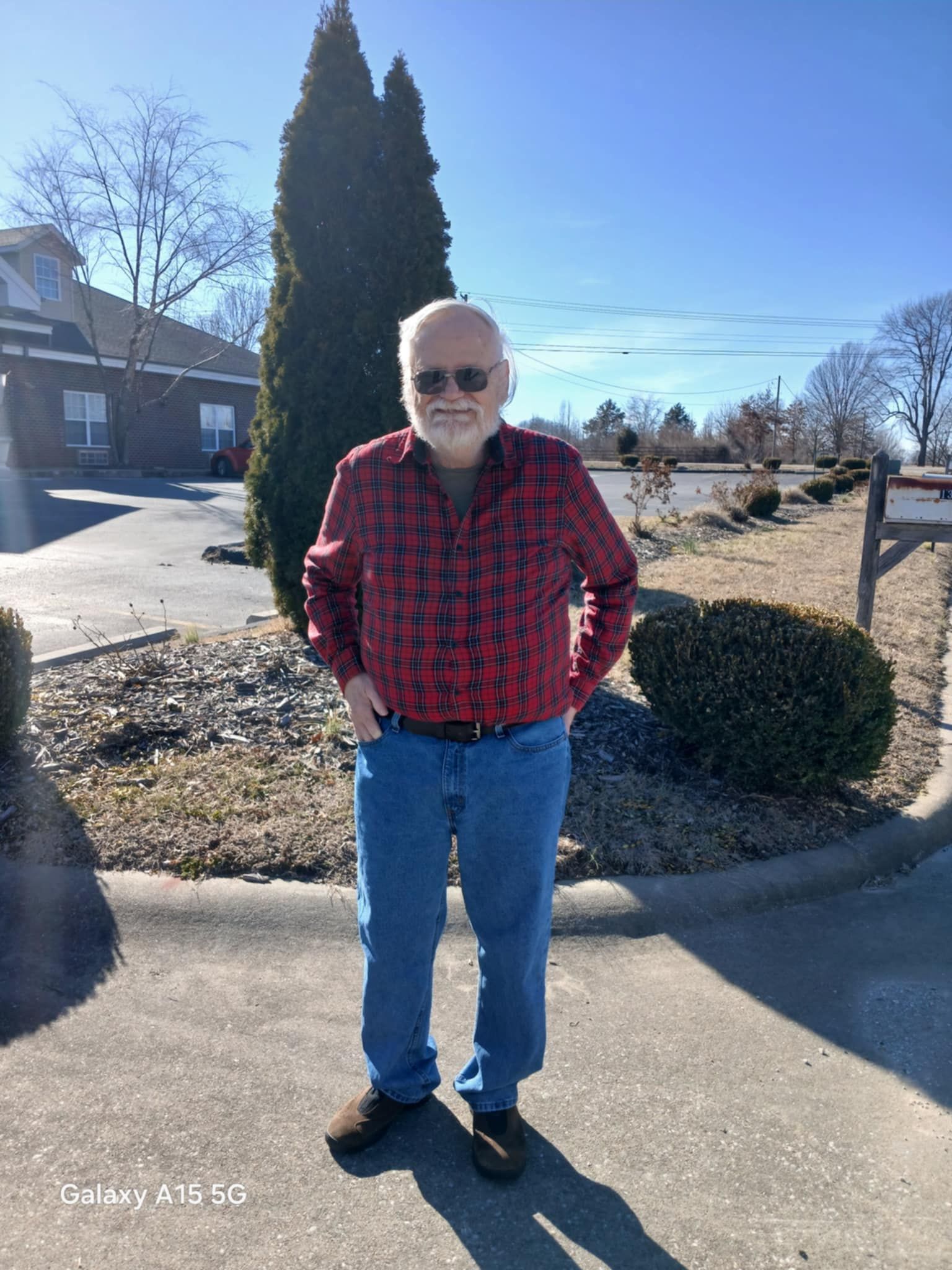A man in a red plaid shirt and blue jeans is standing on a sidewalk.