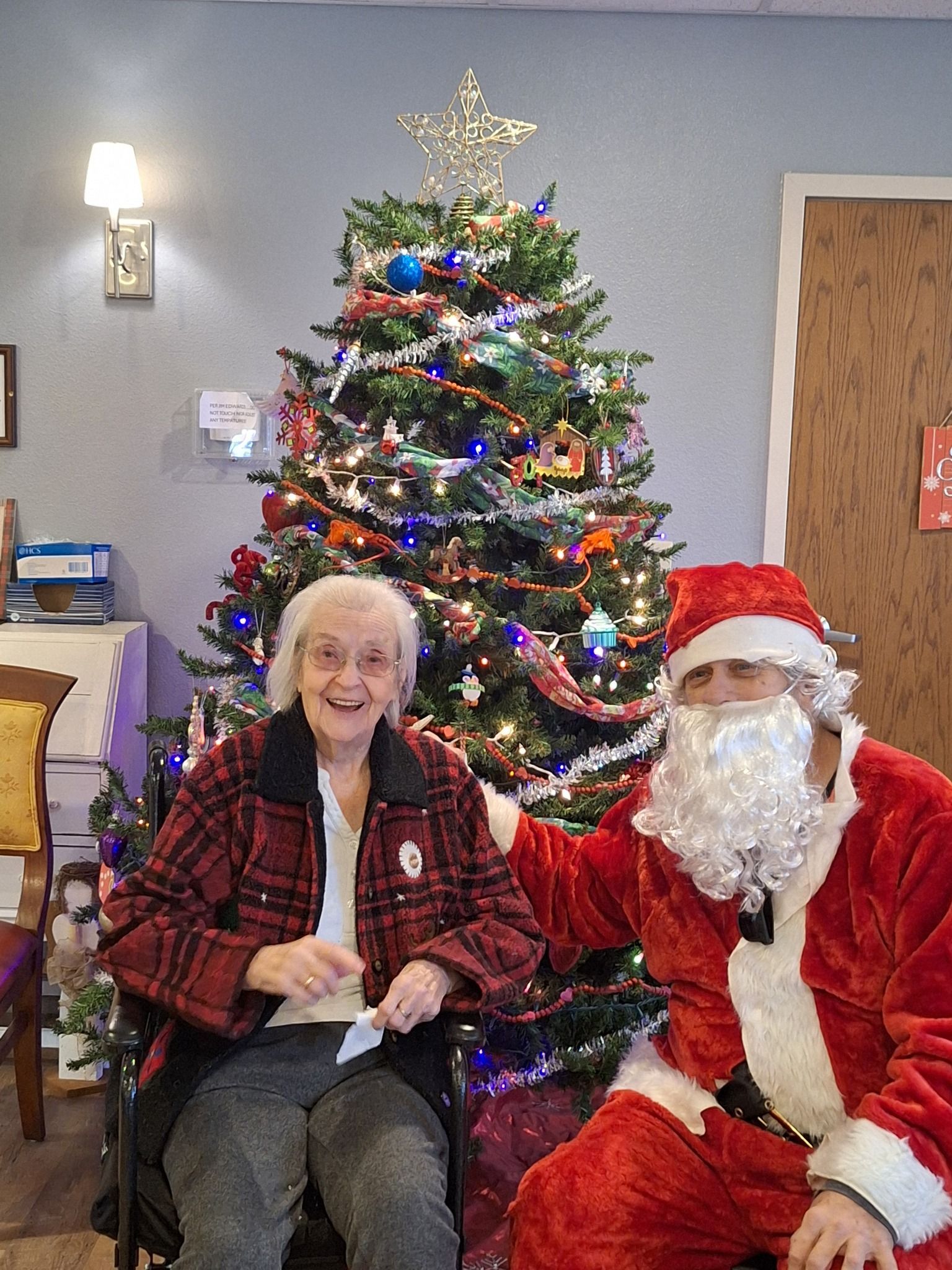 A man dressed as santa claus is sitting next to a woman in a wheelchair in front of a christmas tree.