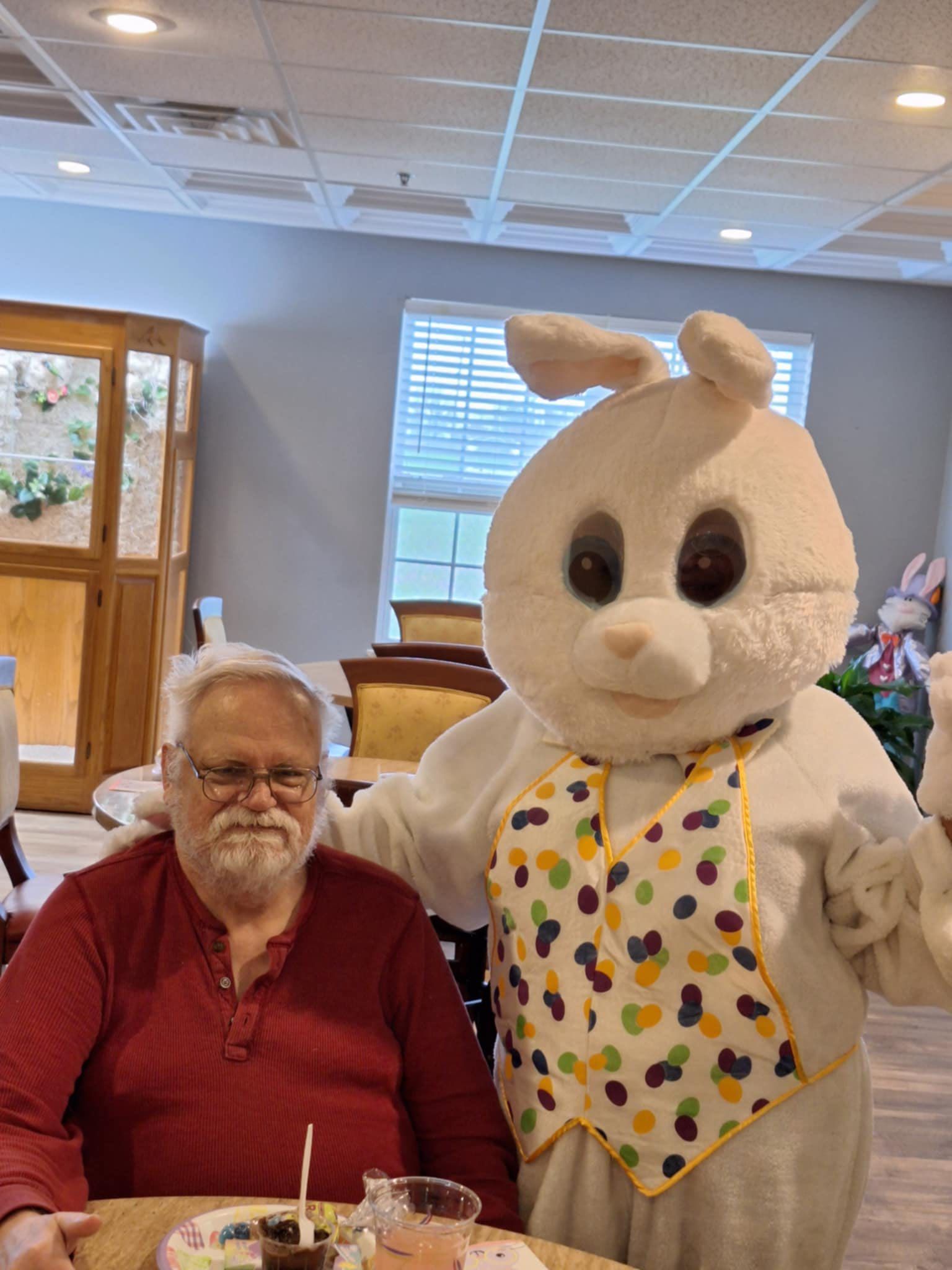 A man in a red shirt is sitting at a table next to a bunny mascot.