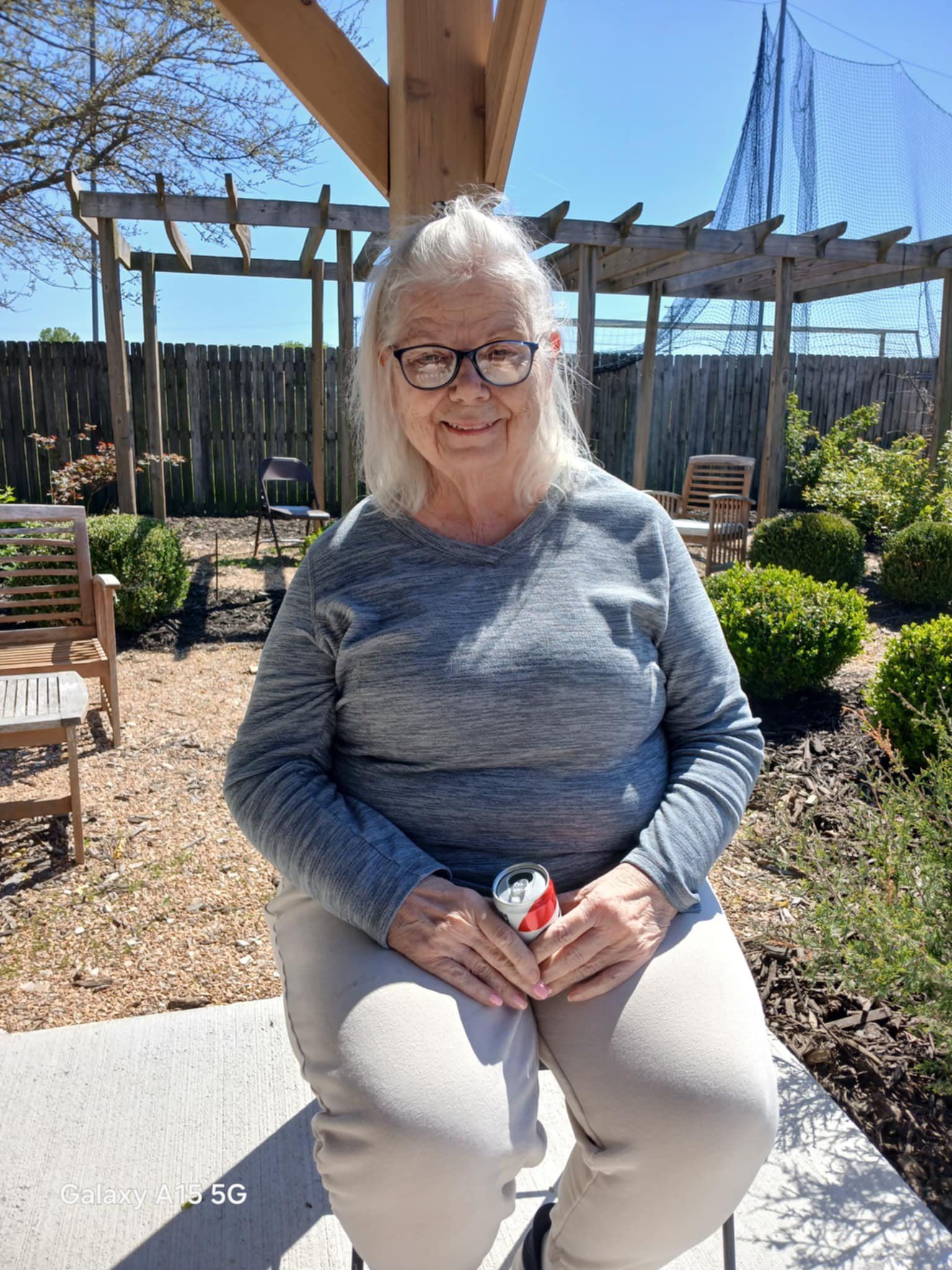 An elderly woman wearing glasses is sitting on a bench holding a can of soda.