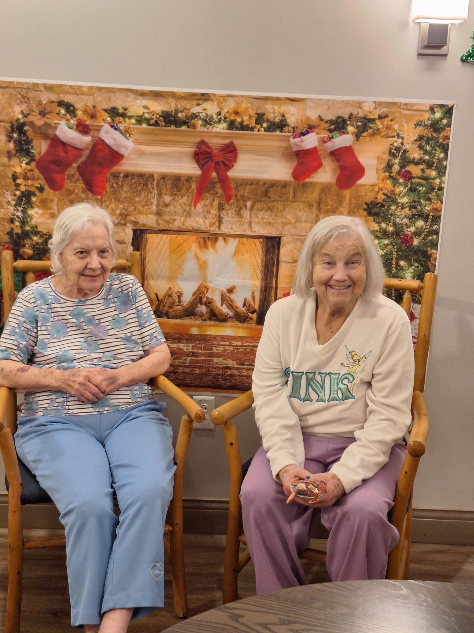 Two elderly women are sitting in chairs in front of a fireplace.