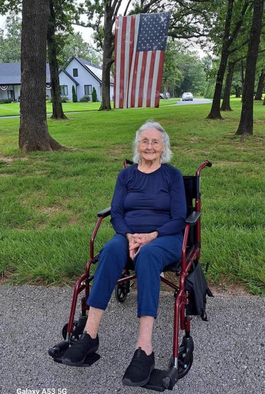 An elderly woman is sitting in a wheelchair in front of an american flag.