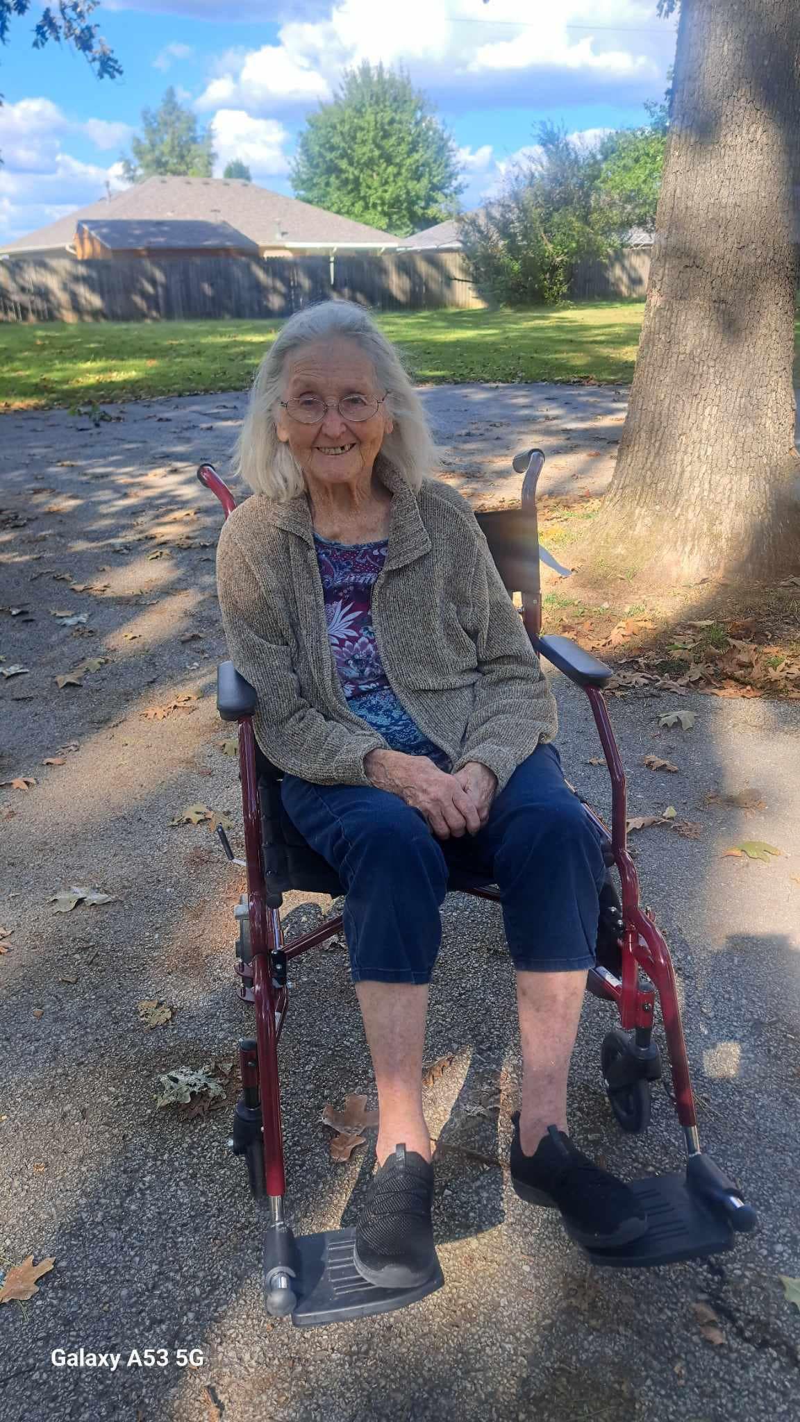 An elderly woman is sitting in a wheelchair in front of a tree.