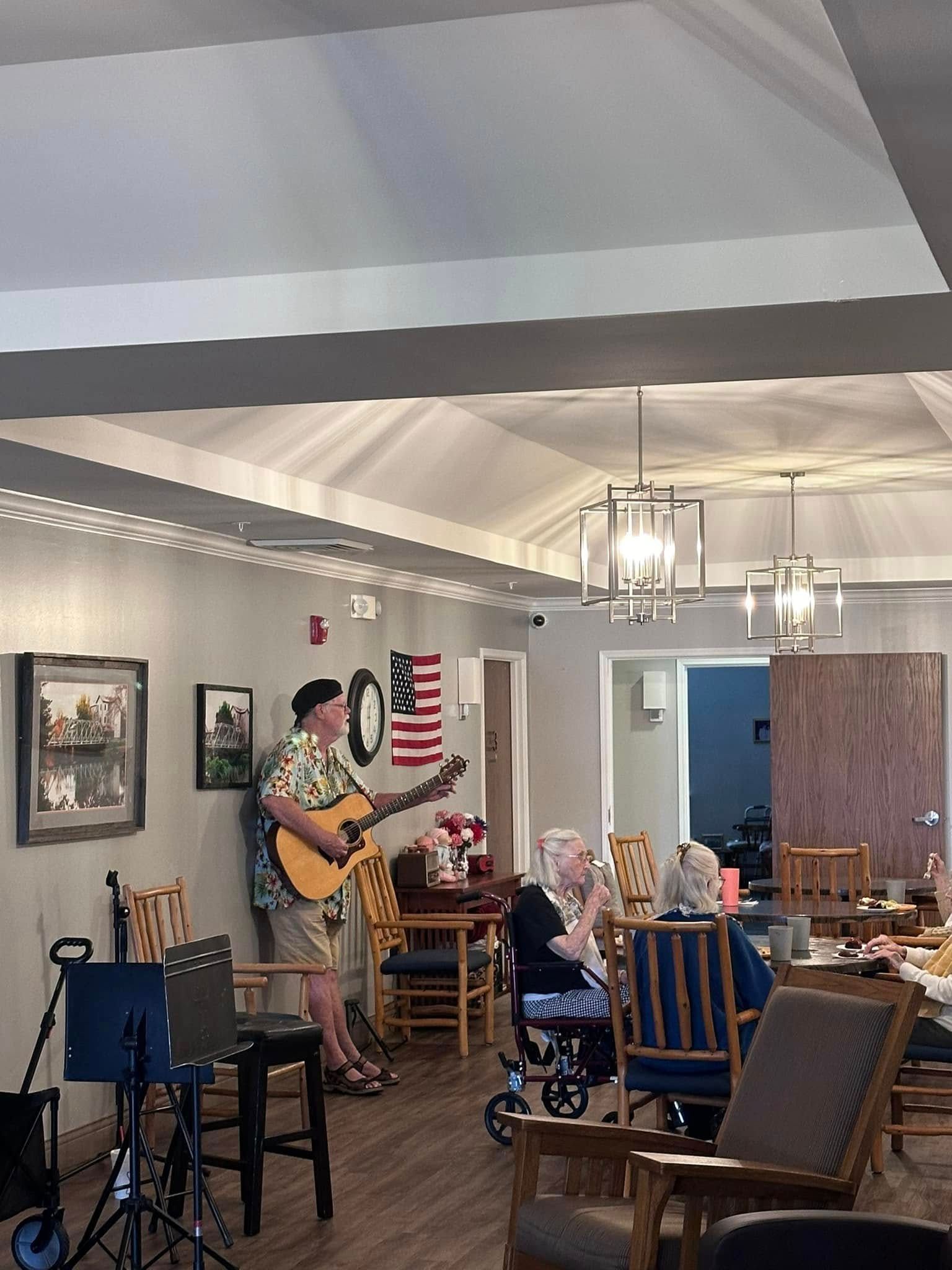 A man is playing a guitar in a room with people sitting in chairs.
