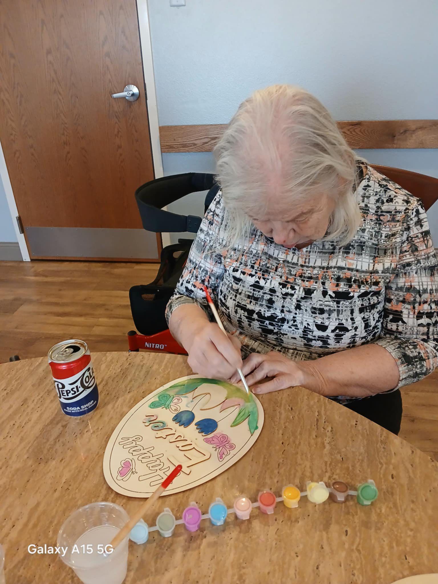 A woman is sitting at a table painting a picture on a piece of wood.
